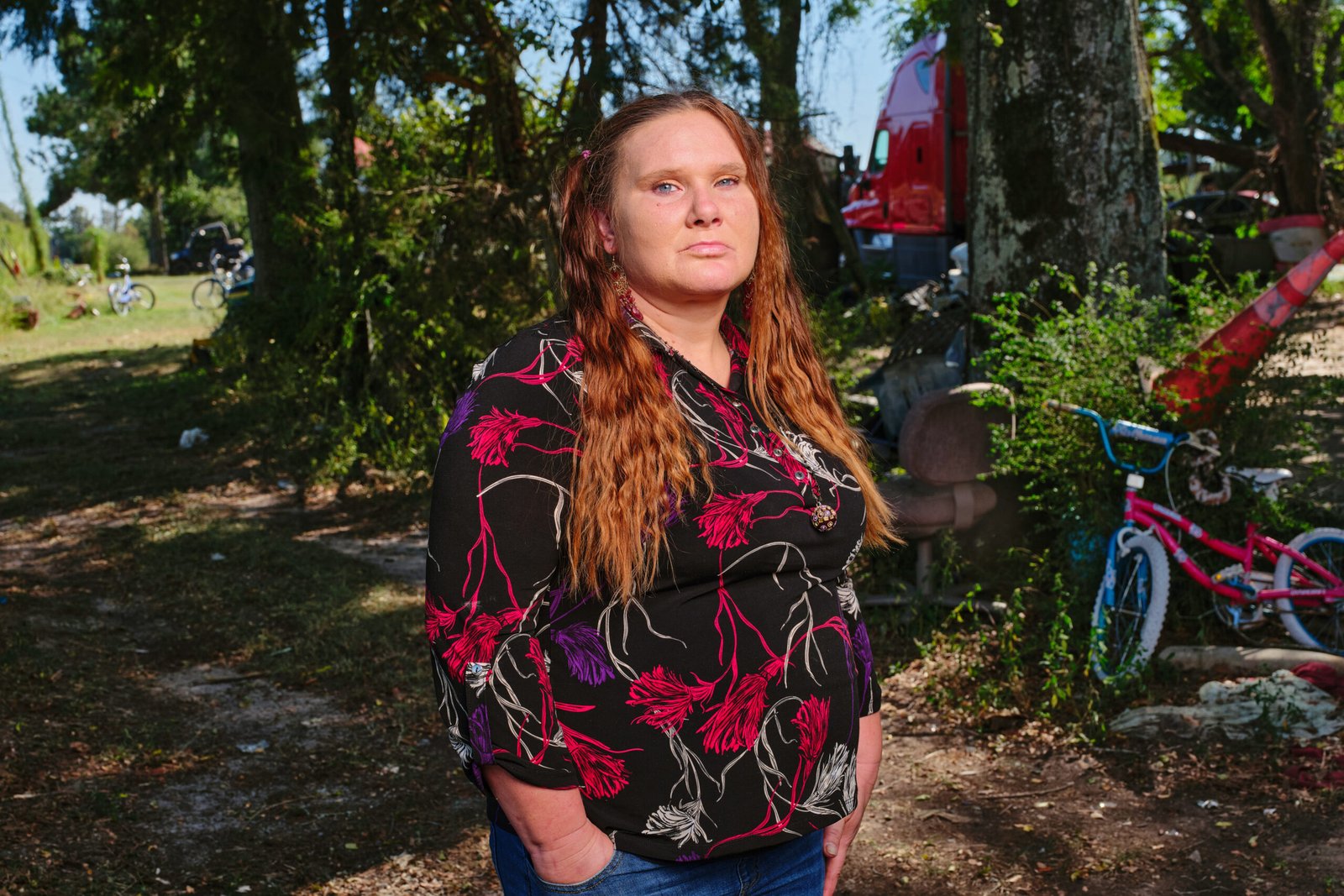 Laura Leigh Taylor stands in a field in Georgia. The front of a truck and a child's bicycle are seen amidst the trees behind her.